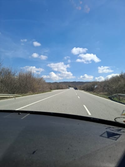 The image shows a two-lane road in Bogyovtsi, Bulgaria, flanked by dry, leafless trees on either side. The sky is blue with scattered white clouds. A vehicle is visible ahead on the road, along with a few other vehicles in the distance. The image is taken from inside a vehicle, with part of the vehicle's interior visible on the right side of the frame.
