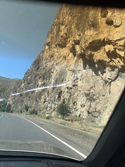 The image shows a view from inside a vehicle traveling along a road next to a rocky cliff face. The road appears to be asphalt with white lane markings. The cliff is steep and has varied textures and colors, ranging from tan and yellow to darker gray and brown. There is some sparse vegetation growing on the cliff and at its base. The sky is clear and blue. The scene is likely located in or near the city of Arpi, Armenia, given the context of the prompt. There are no people or specific actions visible in the image.
