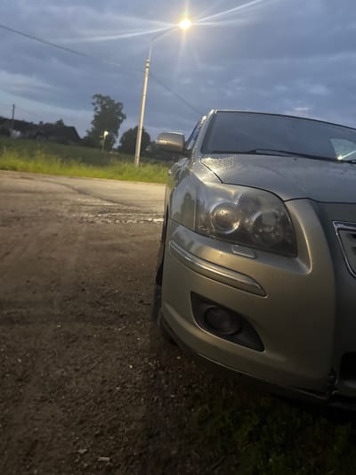 This is a rotated, eye-level, close-up shot of the front of a silver car. The car's headlights and part of the bumper are clearly visible. The lighting suggests it is dusk or dawn, with a streetlamp on the left emitting a bright light that causes lens flare. The sky is a deep blue with some clouds, and the overall atmosphere is dim. The ground appears to be a gravelly or dirt road, with some wetness visible. Green grass and trees are visible in the background, along with a few houses and structures. The car appears to be parked outdoors, possibly in a rural or semi-rural area. There is no text visible in the image.