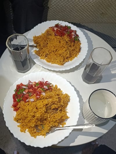 Two plates of rice topped with tomato and onion salsa sit on a round, white table. A fork rests on the lower plate. Next to each plate is a glass, one filled with a dark liquid and the other with a clear one. An empty mug sits beside the glass of clear liquid. The setting suggests a casual dining arrangement indoors.