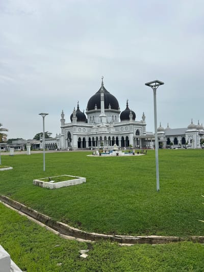 The image shows the exterior of Zahir Mosque in Alor Setar, Malaysia, on an overcast day. The mosque is a large, white building with multiple dark domes and minarets. The main dome is prominent and centrally located. A manicured green lawn surrounds the mosque, with several streetlights and some decorative landscaping visible. In the foreground, there is a drainage ditch and a concrete planter. The overall scene is serene and architectural.