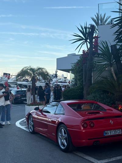 A red sports car, possibly a Ferrari, is parked on a street next to a white building with palm trees. The car has a license plate that reads "MA 9139 CG". Several people are visible in the background, some standing and some seated. They appear to be gathered near a marina, with several boats docked in the distance. The sky is clear and blue, suggesting it is daytime. The scene has a relaxed and upscale atmosphere, possibly a vacation or a social gathering. The time of day appears to be late afternoon or early evening, given the quality of light. The location cues, such as the palm trees and the marina, suggest a coastal area.