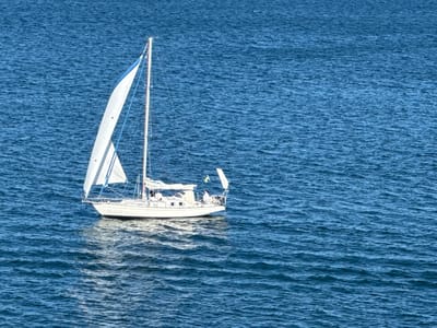 A white sailboat with a blue stripe on its sail is navigating through choppy blue water. The boat has a white hull and a mast with rigging. Two people are visible on the deck of the sailboat. The person at the helm appears to be wearing a white shirt, and another individual is standing near the stern. A small yellow and blue flag is attached to the back of the boat. The water exhibits ripples and reflects the sunlight, creating a textured surface. This scene is likely taking place in the waters surrounding Karlskrona, Sweden, a city known for its naval heritage and archipelago.