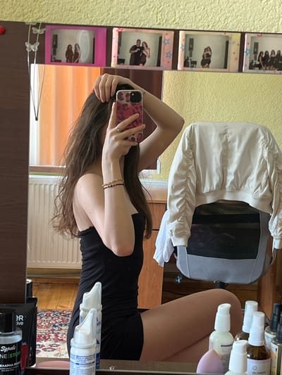 In Popintsi, Bulgaria, a young woman is taking a mirror selfie. She is wearing a black strapless dress and has long, dark, wavy hair. She is holding a pink leopard-print phone in her right hand, with her left hand in her hair. She is wearing two bracelets on her left wrist and a ring on her left index finger.
The background of the image shows a yellow, textured wall with several framed photos mounted above a mirror. The mirror reflects the woman and part of the room, including a radiator, a window with orange curtains, and a wooden dresser. To the right of the woman, a cream-colored jacket is draped over a gray office chair. The foreground shows a vanity surface with various cosmetic bottles and containers. The overall environment appears to be a bedroom or dressing area.