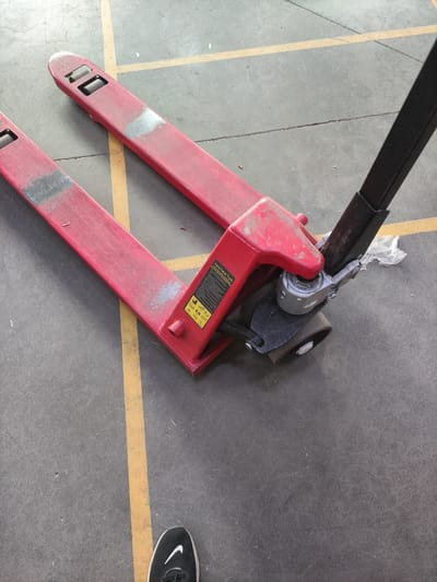 The image shows a red manual pallet jack on a concrete floor. The pallet jack has two long forks that extend forward, and a handle at the rear for steering and lifting. The floor is marked with a yellow line. A person's foot, wearing a black Nike sneaker, is visible in the lower right corner of the image. The scene is likely in an industrial or warehouse setting. There is no information in the image to indicate it is located in Потапово, Russia.