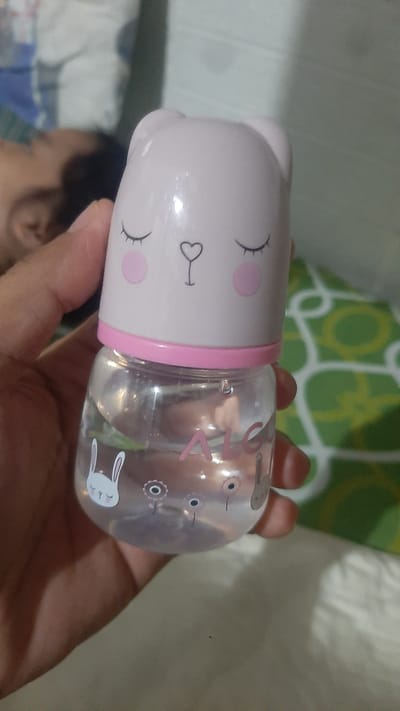 The media file shows a close-up, horizontal shot of a person's hand holding a clear plastic baby bottle with a pink, cartoon-like animal face on one end. The face features closed eyes with eyelashes, two pink circles for cheeks, and a small heart-shaped nose. The bottle itself is filled with a clear liquid. On the bottle's surface, there are decorative elements including pink lettering that appears to say "ALC", and drawings of rabbits and flowers. The hand holding the bottle is partially visible and appears to belong to an adult. In the blurred background, a child's head and shoulder are visible, suggesting the bottle is for a baby. There is also a green and white patterned cushion in the background, and a light-colored surface that could be a bed or couch. The lighting suggests an indoor setting, possibly during the daytime. The overall impression is one of domesticity and childcare. The context provided places this scene in Kawit, Philippines.