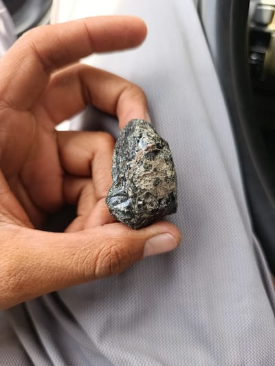 A person's hand, with visible finger creases and skin texture, holds a dark, irregular-shaped object with a rough, textured surface and shiny, glassy areas. The background consists of light grey fabric with a subtle patterned texture and a dark, blurred area on the right, possibly part of a car interior. The scene is lit from above and slightly to the left.