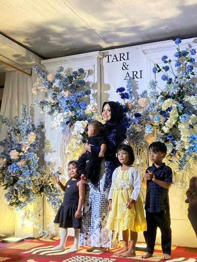 A wedding reception backdrop featuring the names "TARI & ARI" is adorned with large floral arrangements in shades of blue, white, and cream. In front of the backdrop, a woman wearing a dark hijab and a dark blue dress holds a baby. To her left, a young girl in a black polka dot dress and white socks stands with her right arm raised, making a peace sign. To the woman's right, a young girl in a yellow dress and white cardigan stands, followed by a young boy in a blue plaid shirt and black pants. They are positioned on a red and white striped carpeted stage. The scene appears to be indoors, possibly at a venue in Kembang Harum, Indonesia, indicated by a decorative element in the background that suggests a traditional Indonesian wedding setting. Lighting casts shadows on the ceiling, and some of the floral decorations are illuminated.