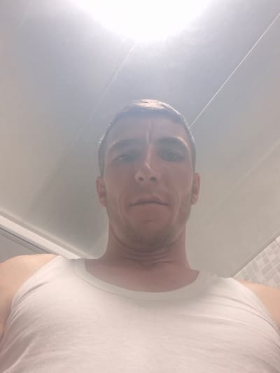 The image is a low-angle, close-up, selfie-style shot of a man's head and upper torso. The man is wearing a white tank top. His face is partially obscured by harsh overhead lighting, creating a bright, washed-out effect, particularly in the upper portion of the image. The background appears to be a ceiling with horizontal paneling. Based on the provided location information, this scene is situated in Hovtamej, Armenia.