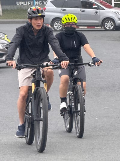 Two individuals are cycling side-by-side on a paved road. The person on the left, a male, wears a black jacket, tan shorts, and a helmet with colorful geometric patterns. He is looking forward and appears to be speaking. The person on the right, a female, wears a black short-sleeved cycling jersey, black cycling shorts, and a bright yellow helmet. She is also wearing glasses and a black face covering. Both are on mountain bikes with front suspension. In the background, a silver SUV is parked. The scene is set on a city street, likely in Da Lat, Vietnam, given the presence of similar SUVs and signage. The road surface is asphalt, and the lighting suggests it is daytime, possibly overcast or late afternoon.