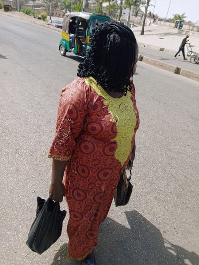 The image captures an outdoor street scene in Mumbwa, Zambia, under bright daylight. The primary subject is an individual, likely female, viewed from the back and left side, walking on an asphalt road. This individual wears a long, reddish-brown dress patterned with orange and maroon concentric circles, featuring a large yellow-green embroidered design on the upper back. Their hair is styled in black braids. They carry a black plastic bag in their right hand and a dark shoulder bag hangs from their left side. A distinct shadow of the person is cast on the road to their right.
In the mid-ground, a green and yellow three-wheeled auto-rickshaw (tuk-tuk) with at least one occupant in a blue shirt is visible on the road, moving in the same general direction. Further in the background on the left, another vehicle, possibly a car, is partially visible.
To the right of the asphalt road, a concrete curb borders a dusty area with sparse vegetation and palm trees. Further down the road on the right, a male figure in dark clothing is pushing a two-wheeled cart. Distant buildings and rising terrain are visible in the background, with the faint outline of another auto-rickshaw far in the distance on the right.