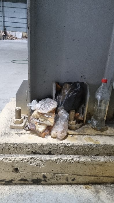 A narrow space is cluttered with trash. A plastic water bottle with a red cap lies near the top. Underneath, there's a black plastic bag and several cellophane-wrapped items, possibly food. The trash is nestled between a gray, metallic structure and a rough concrete wall, where a bolt and nut are visible. A glimpse of a metal fence and a green hose is seen in the background.