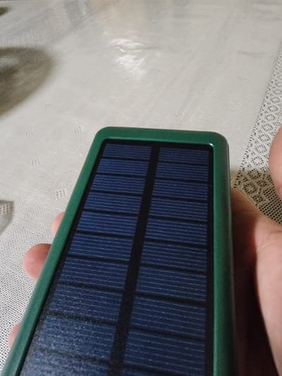 A person's hand is holding a small, green-framed solar panel charger. The solar panel is divided into multiple blue rows with thin white lines. The background is a textured, light-colored surface, possibly a tablecloth. The image is an indoor close-up, and there are no discernible landmarks or contextual clues to suggest a specific location within Gudermes, Russia.