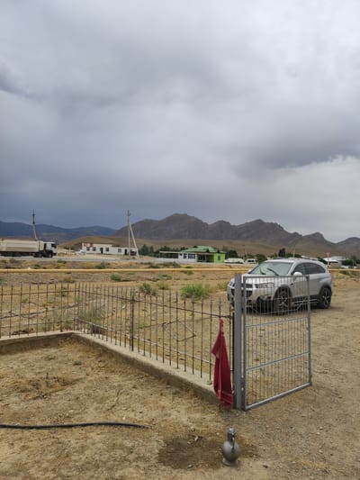 In the city of Egizbulak, Uzbekistan, a grey SUV is parked in a dirt yard. In the foreground, a metal fence surrounds a patch of dry grass and dirt. A red cloth is hanging from the fence, and a metal pitcher sits on the ground. In the background, buildings and mountains can be seen under a cloudy sky. A large white truck is driving on a road in the distance.