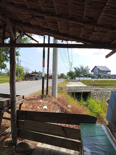 The image depicts a static daytime scene in Keudealuerheung, Indonesia, viewed from within a rudimentary structure featuring a dark thatched roof and wooden support beams in the immediate foreground. The structure's interior on the left includes a wooden table or platform, while the right side shows a surface covered with a green tarp.

Beyond the structure, a paved asphalt road extends horizontally across the midground. The road shoulders are unpaved, covered with dry vegetation, and exhibit scattered refuse, including white plastic bags. Several utility poles stand alongside the road: a prominent light-grey concrete pole centrally positioned, and two thinner poles, one striped orange and black, to its left. Overhead utility lines are visible, stretching across the frame.

To the right of the road, a concrete bridge or culvert spans a narrow, vegetated waterway or ditch. Beyond this, a large, modern multi-story building with a dark grey gabled roof and light-colored walls is visible in the background, situated behind a white fence and a flat, open area, possibly a field. Further down the road on the right, a yellow vehicle is partially discernible, heading away from the observer. On the left side of the road in the background, other smaller structures with dark roofs are present amidst green foliage. The sky is bright blue with scattered white clouds. No individuals are visible.