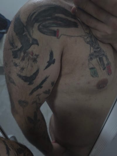 A man's upper body is shown with extensive tattoos. On his chest, there's a tattoo of what appears to be a paint gun. His shoulder and arm are covered with a large piece featuring crows and a brush-like design, extending down his arm. The tattoos have a dark, ink-heavy style. The skin appears to be fair with some texture and hair growth on the arm. The composition is angled, showcasing a close-up perspective of the tattooed areas.