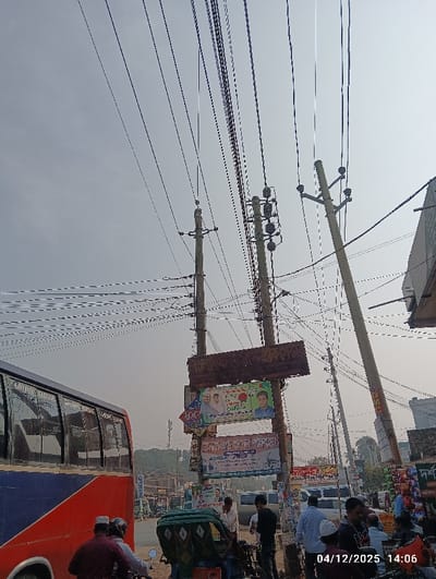 The image depicts an urban street scene in Kaliganj, Bangladesh, recorded at 14:06 on December 4, 2025. The sky is clear, dominated by a dense network of black electrical and communication wires strung between multiple concrete utility poles that extend vertically across the frame. Two prominent poles in the midground are heavily equipped with transformers and insulators. Attached to these poles is a large, weathered, rectangular brown sign with illegible text, beneath which several colorful political and commercial banners in Bengali are displayed.
On the left side, the rear-left portion of a red and blue passenger bus is visible. In the immediate foreground, a man wearing a red long-sleeved shirt and a white skullcap is seated on a motorcycle, facing away from the viewer. Adjacent to him is a green three-wheeled auto-rickshaw. A cluster of approximately ten individuals, predominantly adult males, are congregated on foot near the utility poles and a roadside area on the right, some appearing to interact or observe. Various other vehicles, including cars and additional motorcycles, are visible further down the street. The background features several buildings, distant trees, and a cellular communication tower. The scene indicates a busy commercial or transportation thoroughfare.