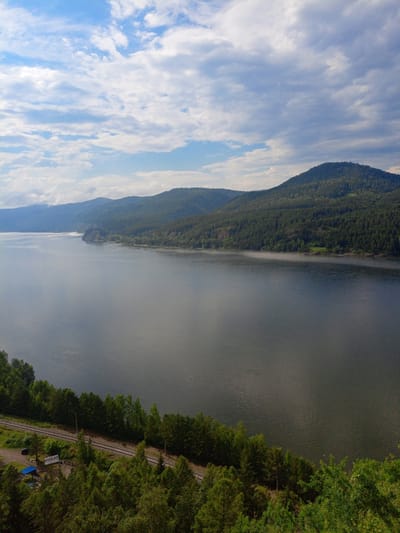 The image shows a wide, calm river flowing through a forested mountain landscape. The sky is partly cloudy with blue sky visible. The mountains are covered in dense green trees, with some lighter green patches suggesting clearings or different types of vegetation. A railway track runs along the near bank of the river, lined by trees. In the foreground, there are more trees, and a small blue structure and a white sign with Russian text are visible near the railway. The river surface is dark and reflects the sky and surrounding mountains. The overall impression is a serene natural scene. This location is identified as Sliznevo, Russia, suggesting a view of the Yenisei River.