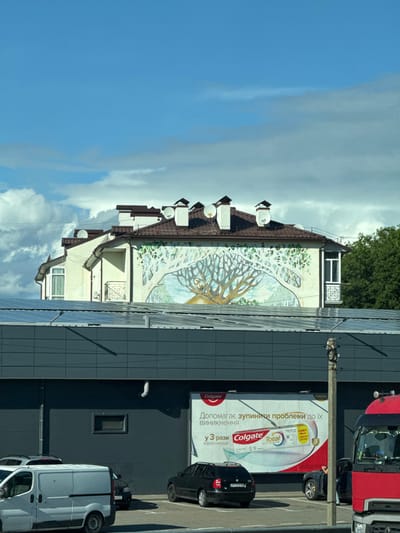 The image depicts an urban scene on Peremohy Street, Ukraine, under a partially cloudy blue sky during daylight. In the foreground, a modern, dark gray building features a flat roof covered with numerous solar panels. On its facade, a large billboard displays an advertisement for Colgate Total toothpaste in Ukrainian. A parking lot is visible in front of this building, occupied by a white cargo van, a black hatchback, another dark vehicle, and the partial front of a red semi-truck on the far right. A concrete utility pole stands in the right midground. Behind the dark gray building, a lighter-colored, multi-story building with a brown-tiled pitched roof is prominent. Its main facade features a large mural of a stylized tree with an owl perched within its branches, against a light blue and green backdrop. The roof of this background building is equipped with multiple brown chimneys and several white satellite dishes. Green trees are visible on the far right, behind the background building. No people are discernible in the scene.
