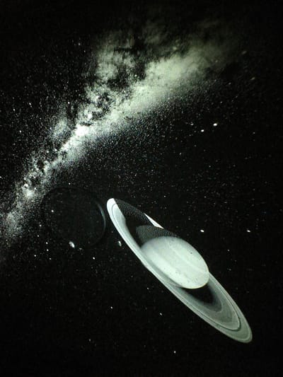 The image depicts a dark, star-filled background, possibly representing space. A planet, resembling Saturn with its distinctive rings, is prominently featured in the lower right quadrant. To the left of the planet, a large, dark, circular object is visible, potentially another celestial body or a feature within the simulated space environment. A nebulous, white cloud-like formation stretches across the upper portion of the image, adding to the cosmic atmosphere. The overall scene appears to be a projection or a simulated display, rather than a photograph of an actual location in Jiguan District, China. There are no people or identifiable structures from the city visible in the image.