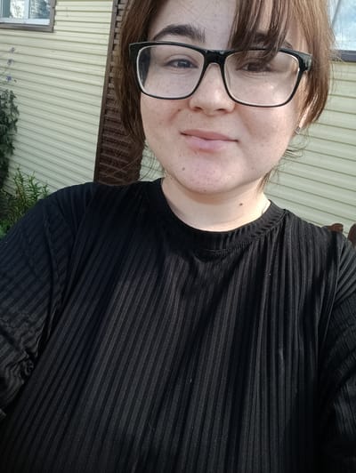 A young woman wearing glasses and a black ribbed t-shirt is photographed from the chest up, outdoors, in front of a light-colored, horizontally-paneled building. The building appears to be a shed or small structure. A window with a dark frame is visible to the left of the woman, and some green foliage is to the far left of the frame. The woman is looking towards the camera with a slight smile. She has light brown hair with bangs that fall across her forehead, and she has freckles on her face. The background is blurred, but the building's siding is clearly visible. The image appears to be taken in a rural or suburban setting.