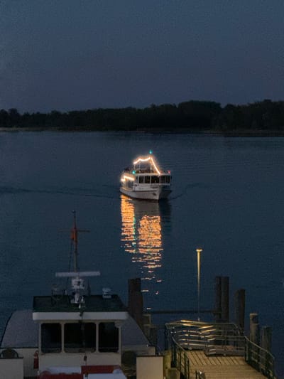 This is a nighttime, eye-level, full-frame shot of a ferry illuminated with lights, cruising on a dark body of water. The ferry is decorated with strings of warm-toned lights that create a reflection on the water. In the foreground, a portion of a dock is visible, with wooden planks and railings. A single lamppost on the dock emits a warm glow. In the background, a silhouetted tree line runs along the horizon. The water is dark and calm. This scene is located on Lake Chiemsee in Prien am Chiemsee, Germany.