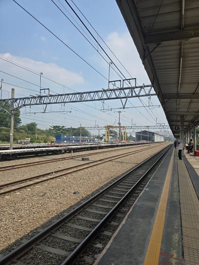 The image depicts an outdoor scene at a railway station in Klapanunggal, Indonesia. The foreground is dominated by parallel train tracks, with gravel ballast between the ties. To the right, a concrete platform runs along the tracks, featuring yellow tactile paving and a low concrete wall. Several people are visible on the platform, including two individuals standing near a red bin and another two walking away from the camera. Overhead, complex electrical infrastructure for the trains, including numerous wires and support structures, spans across the frame. In the middle ground, train cars, some carrying large blue shipping containers, are parked on adjacent tracks. Beyond the tracks, a line of trees forms the horizon, with industrial structures, including a yellow gantry crane and a long, shed-like building, visible in the distance. The sky is predominantly blue with scattered white clouds.
