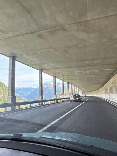 The image shows a view from inside a vehicle traveling on a curved, elevated highway in Airolo, Switzerland. The highway is covered by a concrete structure supported by pillars. To the left, there is a scenic view of a valley with steep, green mountains, a village, and snow-capped peaks in the distance under a cloudy sky. Two cars are visible on the road ahead, with one in the foreground and another further in the distance. The interior of the vehicle, including the dashboard and windshield wipers, is visible in the foreground.