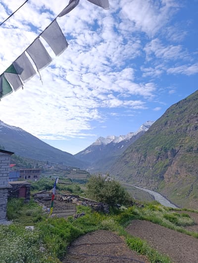 This image captures a majestic mountain landscape with a river flowing through a valley. In the foreground, prayer flags flutter against a bright blue sky dotted with white, fluffy clouds. The flags, predominantly white with some green, are a vibrant symbol against the natural backdrop.
To the left of the frame, a stone building with a red door is visible, suggesting a rural or remote settlement. Further into the valley, a cluster of buildings forms a small town or village nestled amidst terraced fields that climb the hillsides. The fields appear to be cultivated, with some areas tilled and others covered in green vegetation and small white flowers.
The mountains on either side of the valley are rugged and imposing. The slopes are a mix of rocky, barren terrain and areas covered in green vegetation. In the distance, the peaks of the mountains are capped with snow, indicating a high-altitude location. The river, appearing wide and somewhat turbulent, winds its way through the valley, reflecting the sky.
The time of day appears to be morning or early afternoon, given the bright sunlight and the quality of the light. The weather is clear and pleasant, with a visible abundance of clouds. The overall mood of the image is serene and awe-inspiring, highlighting the grandeur of nature and the simple life of the communities that inhabit such remote areas. There is no visible text in the image.