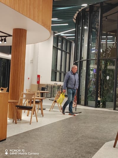 A man wearing a blue puffer jacket and jeans walks across a gray and white tiled floor. He carries a yellow shopping bag in his left hand. To his left, there are wooden chairs and tables near a wooden pillar. Behind him, large windows with black frames reveal greenery inside. The architecture features a curved ceiling detail and modern lighting. A sign is visible to his right. The image includes text indicating it was taken with a Honor 70 phone.