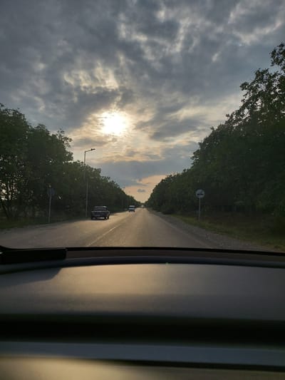 The image, taken from the interior of a vehicle, shows a road lined with dense green trees on both sides. The sun is visible through a cloudy sky, casting a warm glow on the scene. Two cars are traveling on the road, one in the foreground and another further down. There are also streetlights and traffic signs visible along the road. The foreground of the image is dominated by the interior of the car, including the dashboard. The scene depicts a peaceful drive through a natural landscape with a hint of urban infrastructure. Based on the provided metadata, this scene is located in Mirikənd, Azerbaijan.