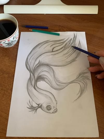 A close-up, overhead shot shows a hand with glittery fingernails holding a dark blue pencil and drawing a sketch of a fish on a white piece of paper. The fish is stylized, with flowing fins that fill most of the paper. The drawing is done in graphite pencil. To the left of the paper, two colored pencils, one green and one blue, lie on a wooden surface. A white object, possibly a clipboard, is also on the left side of the frame. In the bottom left corner, a white ceramic mug with a black and red floral pattern is filled with dark liquid, likely coffee. The scene appears to be set indoors on a wooden table. The lighting suggests it could be daytime. There is no visible text in the image.