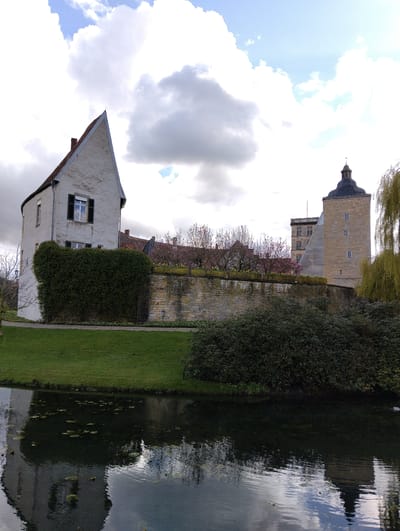 The image depicts a static architectural and natural scene in Burgsteinfurt, Germany, showcasing parts of Steinfurt Castle and its surrounding landscape.
In the foreground, a dark, calm body of water, likely a moat or pond, occupies the lower third of the frame. Its surface reflects the sky and adjacent structures, and several green lily pads are dispersed across it. A strip of vibrant green grass borders the water on the left, leading to a narrow gravel path.
To the left, a multi-story building with white stuccoed walls and a steeply pitched, reddish-brown tiled gable roof stands prominently. A dark chimney is visible on the roofline. The facade features at least one window on the upper floor with dark green louvered shutters. The lower portion of this building's facade is obscured by a dense, manicured green hedge that extends horizontally along a light-colored stone retaining wall.
The stone retaining wall, constructed from irregularly shaped blocks, runs horizontally across the midground, delineating a raised terrace area. Above this wall, on the terrace, several trees display early spring blossoms, including clusters of pink flowers and individual yellow blooms.
On the right side of the image, behind the retaining wall, a substantial square tower of light-colored stone construction rises. It is capped with a dark, multi-tiered, onion-shaped cupola. The tower's facade contains several small, square-paned windows. Behind this tower, sections of another light-colored stone building with an angular roofline are visible. A large weeping willow with delicate light green foliage hangs into the frame from the far right, partially overhanging the water. Dark green shrubs are also present near the water's edge on the right.
The sky is partially overcast, featuring a mix of large white and gray cumulus clouds, with intermittent patches of clear blue sky. No human figures or discernible activity are present in the scene.