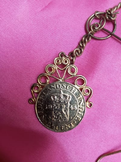 The image displays a metallic charm or pendant resting on a bright pink, subtly textured fabric surface in Qarabucaq qəsəbə, Azerbaijan. The central component of the item is a circular coin, predominantly silver in color but exhibiting a golden sheen due to lighting or patina. The coin's obverse features a prominent crown positioned above a shield emblazoned with a rampant lion. Inscriptions around the coin's perimeter include "NEDERLANDSCH INDIE" at the top, the date "1945" to the left, and "2 1/2 CENT" at the bottom. This coin is encased within an ornate, gold-colored metal filigree frame featuring decorative scrollwork, particularly above the coin. A twisted metal chain or wire extends from the top of the filigree frame upwards and to the right, connecting to a series of intertwined loops and rings, suggesting its attachment to a larger piece of jewelry or an accessory. The pink fabric background shows minor folds and variations in shading. No individuals are present in the scene, and no actions or interactions are depicted.