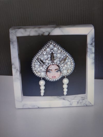 A framed piece of art is displayed against a dark background. The art depicts a portrait of a woman wearing an ornate headdress adorned with pearls and diamonds. The headdress is large and heart-shaped, with dangling pearl earrings. The woman's face is rendered in a stylized manner, with prominent eyes and lips. The frame has a marble-like texture. The background appears to be a wall with horizontal lines. The overall scene is dimly lit, suggesting it might be an indoor setting. No people are present in the immediate vicinity of the artwork. The location in Pospelikha, Russia, is not visually evident from the image.