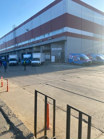 The image displays an exterior view of a large industrial building's loading dock area in Oktyabrskiy, Russia, captured under clear, sunny conditions. The multi-story building features a facade with alternating horizontal dark red and light grey panels. Multiple numbered loading docks are visible along the ground floor, specifically docks 40 through 45, with various roll-up doors, some open, some closed. A prominent white "OZON" logo, accompanied by the Cyrillic word "офис" (office) above it, is affixed to the building's exterior to the right of loading dock 45.

Several vehicles are present on the concrete loading bay surface. Three white box trucks and one blue box truck are parked or positioned near loading docks 40 through 42. To the right, two blue commercial vans, both marked with "OZON" branding and Cyrillic text, are parked near the building's entrance.

Five individuals are visible in the scene. Four individuals are walking away from the viewer on the left side of the frame, one wearing a blue jacket or vest. A fifth individual, wearing a high-visibility yellow-green vest, stands centrally near the parked box trucks, facing away from the camera.

In the immediate foreground, a dark metal railing partially obstructs the lower view. A concrete curb runs along the left side, featuring three orange and white striped bollards. Ground debris is visible along this curb.
