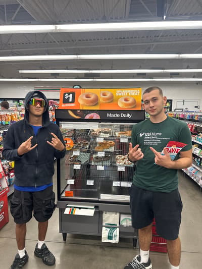 This media file depicts two young men posing inside a brightly lit convenience store, likely a 7-Eleven, in Homosassa Springs, United States of America. The image is oriented sideways, but if viewed upright, shows them standing in front of a donut display.

The man on the right, wearing a dark green t-shirt and black shorts, holds a red bag of "DAVID" brand peanuts and makes a "rock on" hand gesture with both hands. His shirt prominently displays "USFMuma College of Business School of Information Systems and Management UNIVERSITY of SOUTH FLORIDA MS IN BUSINESS ANALYTICS & INFORMATION SYSTEMS". He is also wearing white socks and black and white athletic shoes.

The man on the left, clad in a black hooded sweatshirt, black shorts, and mirrored sunglasses, also makes a similar hand gesture. A blue shirt is visible under his hoodie, and he wears white socks and black athletic shoes.

Behind them is a tiered display stand showcasing various donuts, including "GLAZED DONUT" for $1, as well as Apple, Celebration, Cream Cheese, and Plain Cake donuts priced between $1.69 and $1.99. A large yellow sign next to the display reads "YOU DESERVE A SWEET TREAT" and "Made Daily". Shelves filled with other packaged goods and drinks are visible in the background, typical of a retail store setting. Fluorescent lights illuminate the ceiling. Other visible text includes "CAUTION TIP HAZARD" on the display, and "WHAT'S YOUR BEEF?" on a red sign at the bottom. The men appear to be casually posing for a photo.