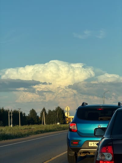 A traffic jam is observed on a paved road in Derzhavino, Russia. A bright turquoise car, license plate ending in Y11, is stationary in the right lane, with its brake lights illuminated. The rear of a dark-colored car is visible to the right of the turquoise car. The road is bordered by a grassy field and a line of trees. In the distance, several power poles are present. The sky is a clear blue, with large, billowy white clouds dominating the upper half of the frame. The lighting suggests it is late afternoon or early evening, with the sun casting a warm glow on the clouds.