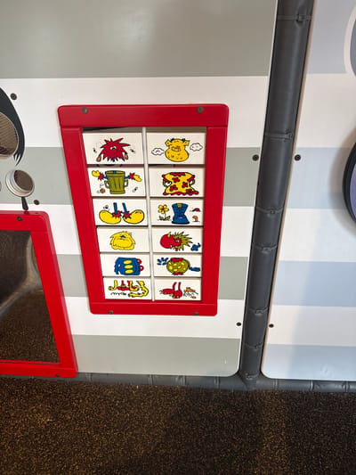 The image shows a detail of a children's play area, likely indoors, with a striped white and gray wall. A red frame holds twelve square tiles, each displaying a cartoon-like image, arranged in two rows. Below this, a partially visible red frame surrounds what appears to be a mirror or another play element. A padded, gray, cylindrical safety barrier runs horizontally above the red-framed tiles. The floor on the right is textured and dark, possibly carpeted. The location is possibly Stone Mountain, United States of America. There are no people visible in the image. The focus is on the play equipment and its details.