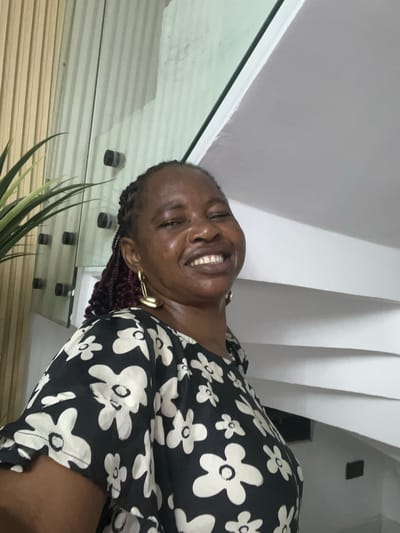 A woman with dark skin, wearing a black and white floral print dress and gold hoop earrings, is smiling broadly. Her hair is styled in braids, with some red strands visible. She is positioned indoors, as indicated by the wall and window in the background. There is a plant in the foreground. It is impossible to determine from the image alone if the location is in Mopo-Akinlade, Nigeria.