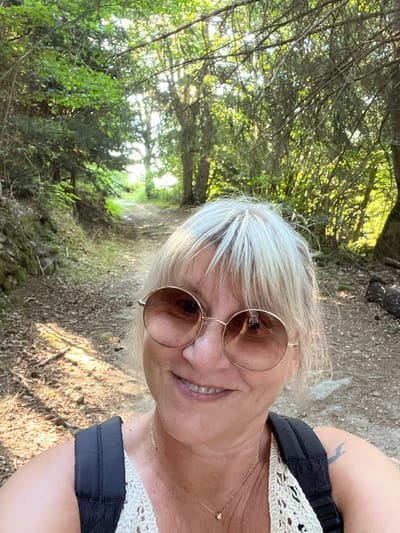 A woman with blonde hair and sunglasses is taking a selfie on a dirt path in a forest. She is smiling and wearing a white crocheted top and a backpack. The sun is shining through the trees, casting dappled shadows on the path. The path appears to be in a rural area, possibly a hiking trail. The woman has a tattoo of a bird on her left shoulder. The image appears to have been taken in Gumières, France.