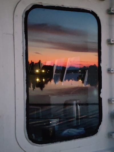 The image shows a reflection in a porthole window on a vessel, likely a ship or boat. The reflection captures a sunset over a body of water, with colorful clouds in hues of orange, pink, and blue dominating the sky. Silhouetted trees and structures are visible along the shoreline, along with several illuminated lights that appear to be streetlights or lights from buildings. The water reflects the sky and the lights, creating a mirror image. The porthole frame is white and shows signs of wear and tear, with some chipped paint. The interior of the vessel is partially visible through the lower part of the window, showing dark surfaces and some indistinct objects. There is no clear indication of the city of Konakovo, Russia, within the image itself, but it is stated as the location.