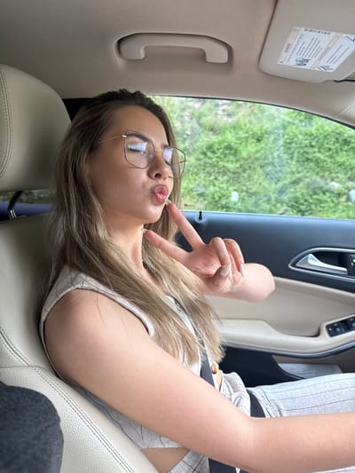 Here's a concise description of the image:
A young woman wearing glasses and a light-colored top is reclining in the passenger seat of a car. She's making a peace sign with her hand. The car interior is light-colored, with the window showing a blurred view of green foliage. A warning label is visible on the sun visor. There is no immediate indication that this picture was taken in Pastra, Bulgaria.