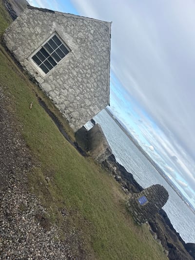 The scene depicts a coastal landscape in Montrose, United Kingdom, under an overcast sky. A prominent, single-story building constructed of grey, rough-hewn stone occupies the left side of the frame. It features one visible, rectangular window with a white multi-paned frame. The building stands on a gently sloping area of green grass, which transitions into a dark gravel path in the foreground.

Adjacent to the building and extending toward the water is a segment of a weathered, grey stone wall. The water itself, a wide body appearing dark blue-grey and rippled, defines the middle ground and background, suggesting an estuary or open sea. The immediate shoreline consists of dark, rugged rocks.

Further along the rocky shore, to the right and closer to the water, a distinctive conical stone monument or cairn is positioned. This structure incorporates a small, square blue plaque on its visible face. The horizon line is visible across the water, with a band of lighter blue sky beneath a broad expanse of grey clouds. No individuals are present in the image.