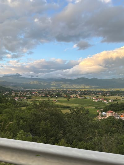 This media file captures a panoramic, slightly rotated view of a scenic valley in Miločani, Montenegro, taken from an elevated perspective, possibly from a road overlooking the area.

In the foreground, a light-colored guardrail is prominent on the right side, partially obscuring a dense cluster of green trees and foliage that fills the lower and right portions of the image.

The midground reveals a vast valley stretching across the landscape. It is dotted with numerous scattered houses and buildings, many featuring red or orange roofs, nestled among a mosaic of agricultural fields and green patches. The settlement appears rural or semi-rural. Further into the valley, cultivated fields are discernible, suggesting agricultural activity.

In the background, a majestic mountain range dominates the horizon. Some mountain peaks are partially enveloped in mist or low clouds. The sky above is a mix of blue and clouds. Bright, golden-tinged clouds suggest either late afternoon or early evening light, contrasting with darker grey clouds scattered throughout the sky.

The scene conveys a tranquil atmosphere. There are no people visible, and no specific activities are taking place. No discernible text is present in the image.