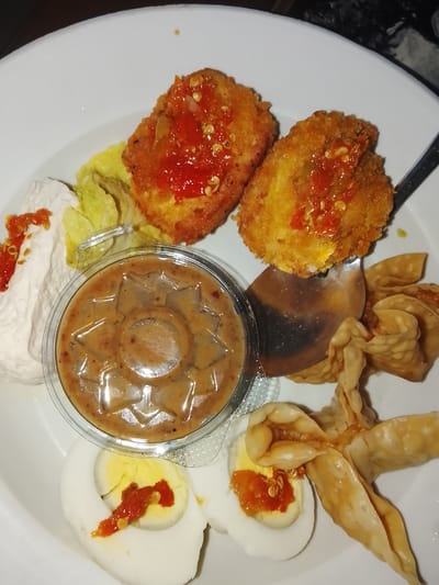 A close-up, overhead shot shows a white plate of Indonesian food. Two golden-brown, crispy fried items, likely tempura or fritters, are topped with a red chili sauce. To their left is a small mound of white tofu or cheese with a drizzle of red sauce, and a few leaves of green lettuce. In the center, a small, clear plastic container holds a thick, brown peanut sauce with visible spices. To the right of the sauce are two fried dumplings, resembling wontons, in a star-like shape. Below these are two halves of a hard-boiled egg, each topped with a dollop of red chili sauce. The background is out of focus, showing a dark, speckled surface, possibly a table. The scene is likely a restaurant or home dining setting in Gunung Malang, Indonesia.