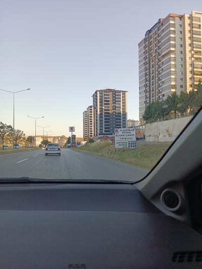 This media file captures a view from inside a vehicle driving on a highway in Kırıkkale, Turkey. The scene is set during the day, with clear skies and sunlight illuminating the landscape. On the right side of the road, a tall, modern apartment building with a glass facade stands prominently. Next to it, a gas station with a Kadell sign is visible, displaying prices for Motorin (diesel), Benzin (gasoline), and LPG. The text on the gas station sign includes "YILDIRIMLAR PETROL ELVAN ÇAKIR" and "TAŞIKO ÇEKLERİNE AKARYAKIT VERILIR".

The highway has multiple lanes, and a white car with the license plate "21 JAE 421" is in front of the viewing vehicle. In the distance, more apartment buildings and residential areas are nestled on a hill. Streetlights line the side of the road, indicating an urban or suburban setting. The overall impression is one of a typical drive through a Turkish city or its outskirts, with modern infrastructure and everyday services readily available. The time of day appears to be late afternoon or early evening, given the angle of the sunlight and the soft, warm light casting long shadows. The weather is fair and clear.