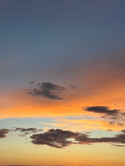 The image captures a dramatic sunset over a rural landscape in Mazeykaŭski Sel'ski Savet, Belarus. The sky is a canvas of vibrant colors, transitioning from a deep blue on the left to fiery oranges, soft yellows, and pale blues on the right, indicative of the twilight hour. Wispy clouds are scattered across the sky, some catching the last rays of the sun and appearing in shades of dark gray, fiery orange, and white. The overall mood is one of awe and the sublime beauty of nature. There are no people or man-made objects visible in the frame, emphasizing the vastness and natural splendor of the scene. The image appears to be a vertical shot, adding to the sense of height and the immensity of the sky.
