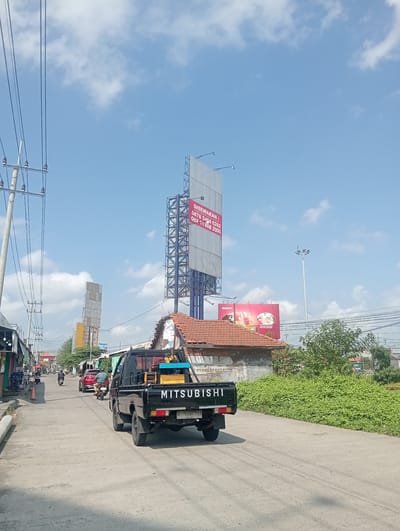 The image depicts a street scene in Sawotratap, Indonesia, under a partly cloudy blue sky. The foreground is dominated by the rear of a black Mitsubishi pickup truck, which is facing away from the camera and driving down a paved road. The truck has a license plate that reads "W 8894 YA". On the bed of the truck, a yellow and blue generator or machinery is visible.
The road is lined with various structures. On the left side, there are several low-rise buildings that appear to be shops or businesses, with some signage visible. Further down the road, traffic is light, with a few motorcycles and a red car also traveling in the same direction as the pickup truck.
In the background, a tall metal advertising billboard structure stands prominently, with some text on a large white board that appears to be a rental advertisement. Another, shorter billboard with a red background and images of food items is also visible. A tall lamppost with multiple lights is situated in the distance, and lush green vegetation lines the right side of the road. Electrical wires and utility poles are present along the left side of the street. The overall impression is a typical urban streetscape in Indonesia.