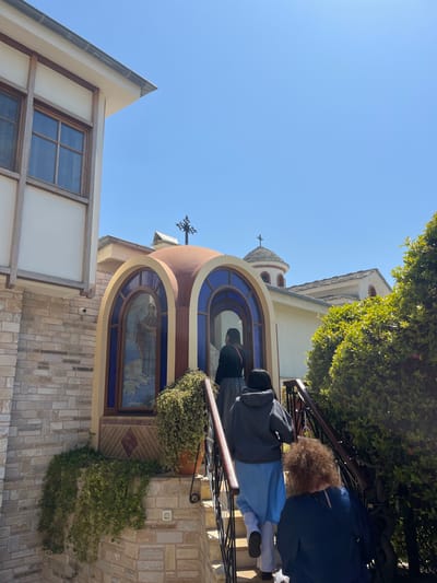 The image shows a small, arched structure with two stained glass windows. The windows depict religious figures. The structure has a red-brown dome and is attached to a larger building, likely a church. The scene takes place in the Moni Archaggelou monastery on the Greek island of Tinos, at the coordinates provided. Four people walk up a set of stone steps to the structure. They are dressed in casual clothing, and one is carrying a bag. The steps are flanked by railings, and a lush green bush grows in the foreground. The building is made of whitewashed stone and has a tiled roof. The sky is clear blue, and the day is sunny.